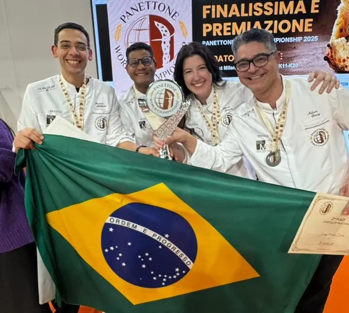 Equipe brasileira liderada pelo padeiro cearense Brunno Malheiros celebra o prêmio de segundo melhor panetone de chocolate do mundo na Panettone World Championship, na Itália, segurando troféu e bandeira do Brasil.