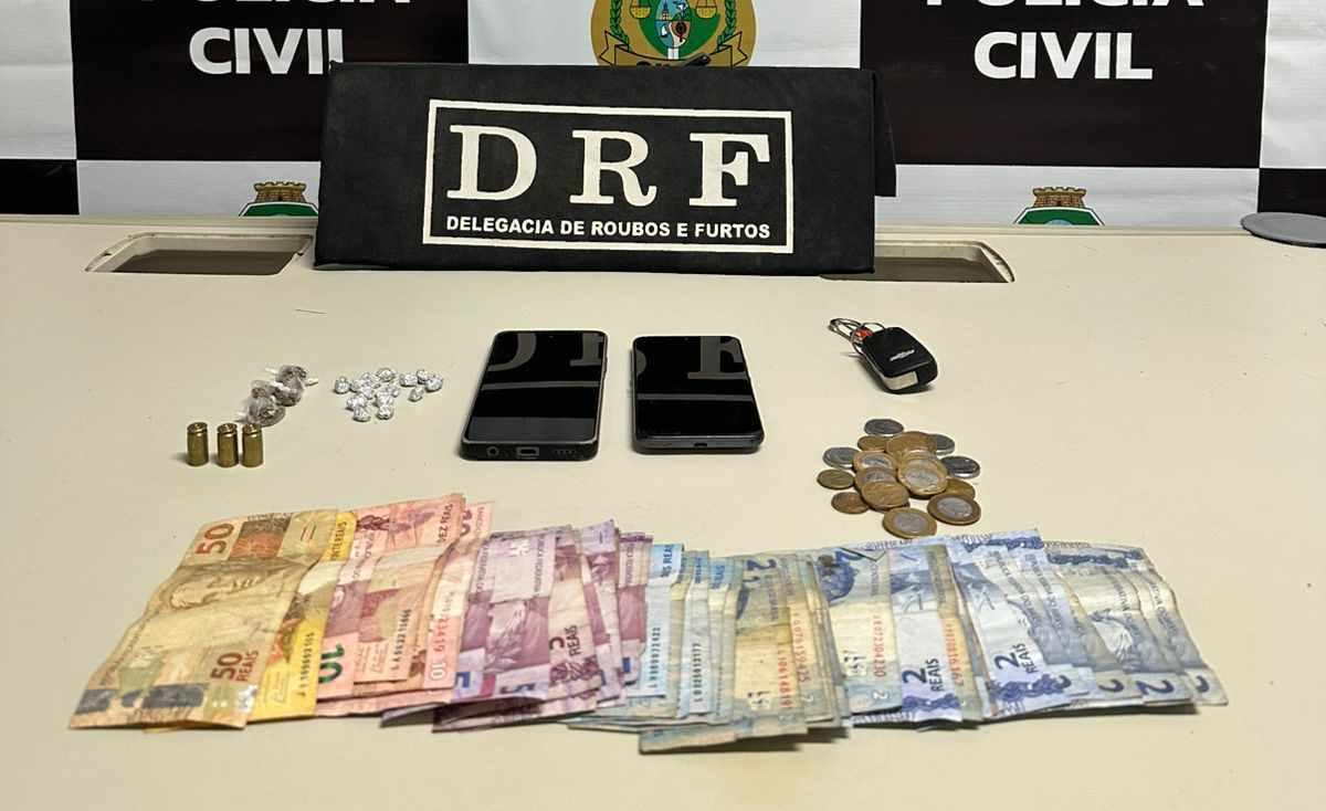 Itens apreendidos pela polícia civil, incluindo dinheiro, dois celulares, chaves, roupas, drogas e um recibo de furto, exibidos em uma mesa com o logotipo da Delegacia de Roubos e Furtos (DRF).