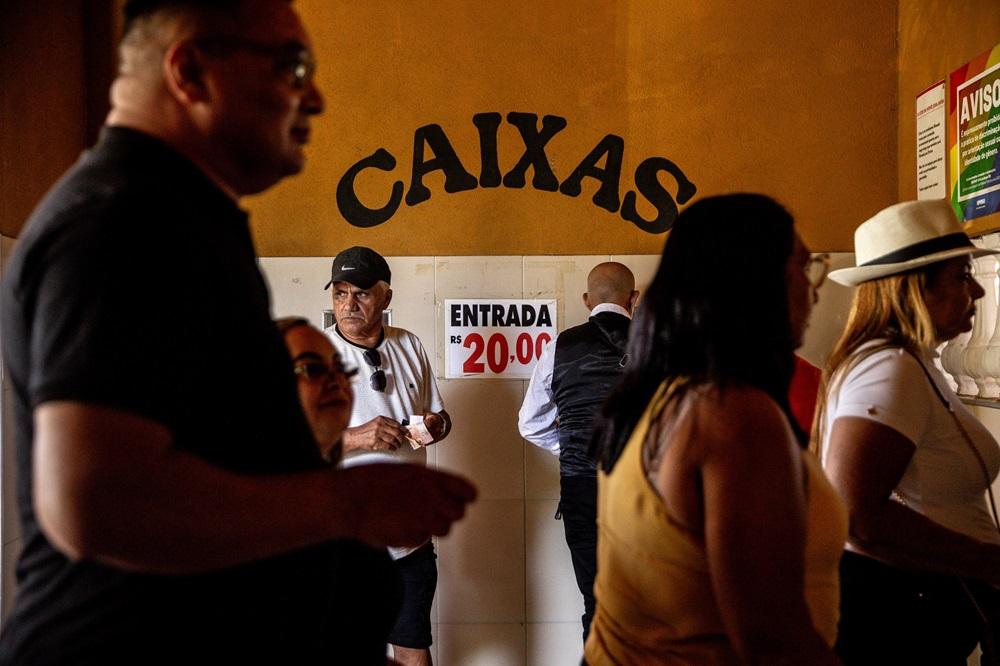 Na imagem, uma fotografia de pessoas em uma área de bilheteria/entrada de um local, com a parede de fundo de cor amarela-mostarda. A palavra 