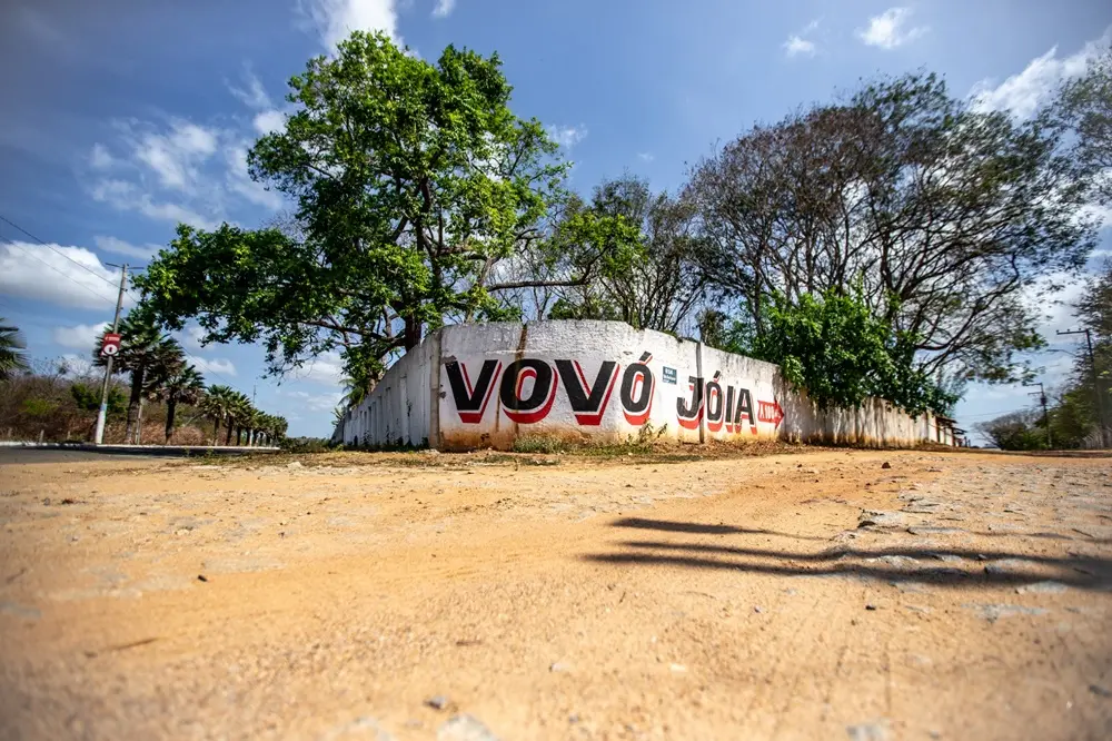 Na imagem, uma imagem em ângulo baixo de um muro branco de concreto em forma de curva em um ambiente rural ou semi-urbano, com a inscrição 
