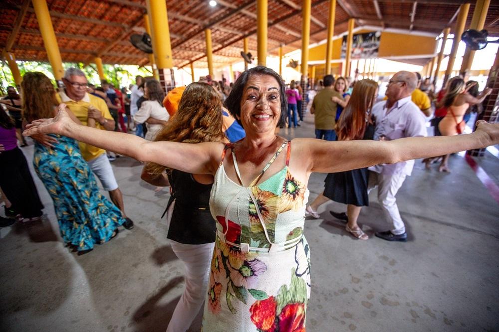 Na imagem, uma mulher sorridente, de meia-idade ou idosa, de pele clara e cabelo escuro, está no centro, em primeiro plano, com os braços esticados, abertos. Ela usa um vestido branco com alças, estampado com grandes flores em tons de amarelo, laranja e verde. Ela está em um salão de festas grande, com teto de telhas e vigas de madeira e pilares amarelos, onde várias outras pessoas dançam em pares ao fundo, a maioria embaçadas, sugerindo movimento. O piso é de concreto cinza. A luz natural brilha no fundo.