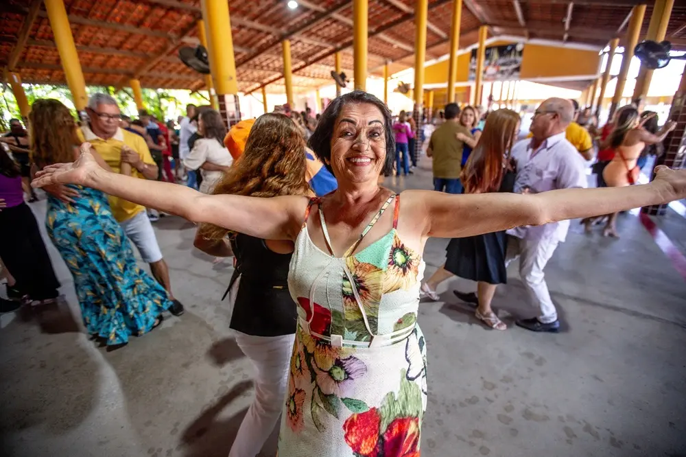 Na imagem, uma mulher sorridente, de meia-idade ou idosa, de pele clara e cabelo escuro, está no centro, em primeiro plano, com os braços esticados, abertos. Ela usa um vestido branco com alças, estampado com grandes flores em tons de amarelo, laranja e verde. Ela está em um salão de festas grande, com teto de telhas e vigas de madeira e pilares amarelos, onde várias outras pessoas dançam em pares ao fundo, a maioria embaçadas, sugerindo movimento. O piso é de concreto cinza. A luz natural brilha no fundo.