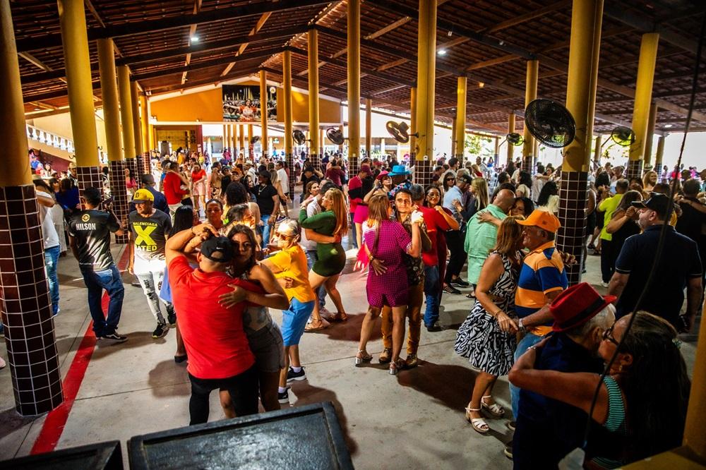 Na imagem, uma visão panorâmica de um grande salão de festas ou evento, lotado de pessoas de várias idades dançando e socializando. O local tem um teto alto com estrutura de madeira e telhas, suportado por numerosos pilares cilíndricos de cor amarela intensa, que criam um corredor ou nave central. As pessoas estão agrupadas em pares e grupos menores no chão de concreto, que tem uma faixa vermelha pintada perto de onde a foto foi tirada. Há ventiladores de parede pendurados em alguns pilares. A iluminação é forte, misturando luz natural e artificial. Muitas pessoas estão abraçadas, dançando forró ou outro ritmo popular. As vestimentas são casuais e coloridas.