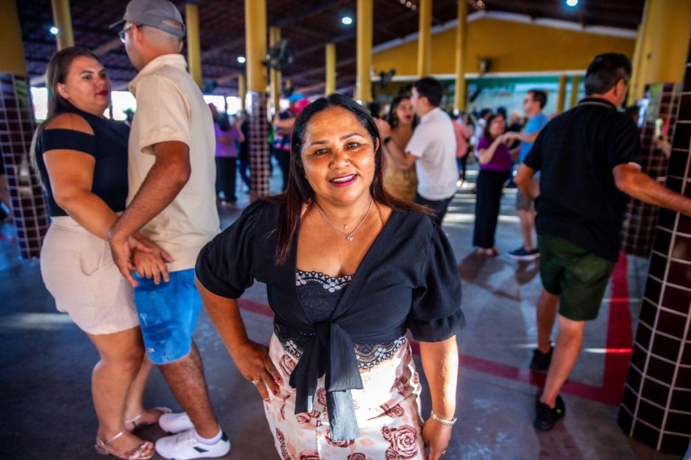 Na imagem, uma mulher sorridente, de pele morena e cabelos escuros, está em primeiro plano e no centro, olhando diretamente para a câmera. Ela usa uma blusa preta de amarração frontal sobre um top de renda preta e uma saia clara com estampa floral em tons de rosa. A mulher está com a mão direita na cintura. Atrás dela, o ambiente é um salão de festas iluminado, com pilares amarelos e teto de madeira. À sua esquerda, um casal dança, com a mulher vestindo uma blusa preta ombro a ombro e saia bege e o homem, uma camisa polo bege e shorts jeans azuis. Outras pessoas estão dançando em segundo plano. O piso é de concreto, com linhas vermelhas e um pilar xadrez de azulejos marrons à direita.