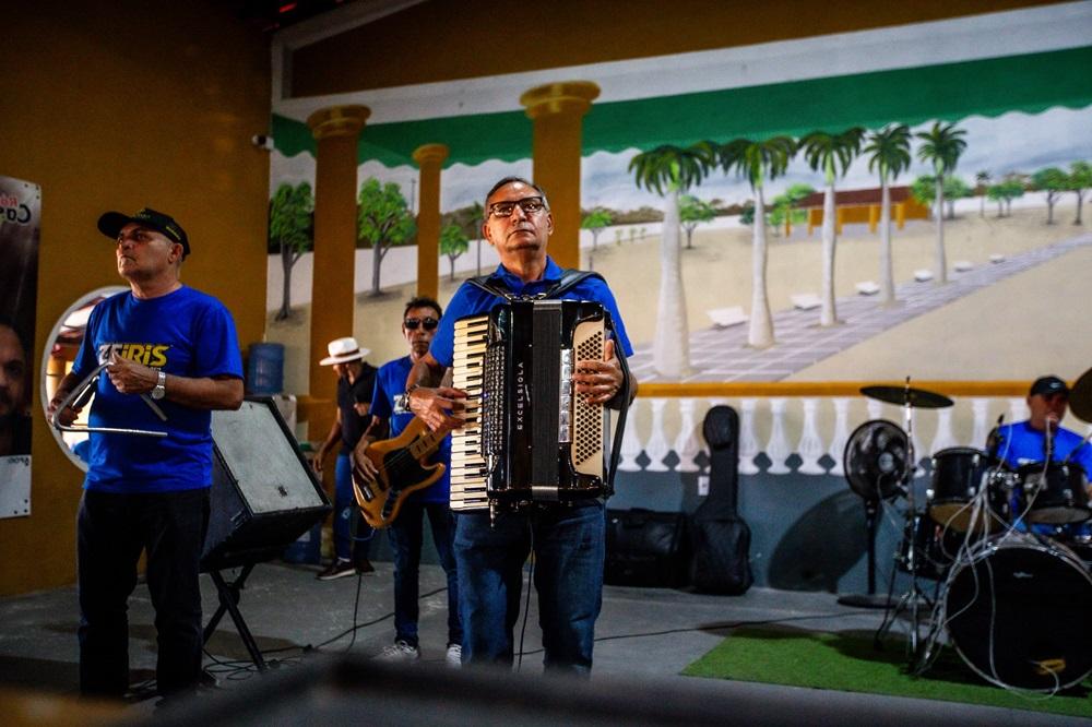 Na imagem, uma banda de forró ou música regional se apresenta em um palco em um ambiente interno. No centro, um homem de pele morena, óculos, camiseta azul e jeans escuros toca um acordeão preto e branco, olhando para cima e para a frente. À sua esquerda, um homem com um boné preto e camiseta azul toca um triângulo. Outro músico com óculos escuros e camiseta azul toca um baixo elétrico amarelo mais ao fundo. À direita, parte de uma bateria preta e um baterista em camiseta azul são visíveis. O fundo do palco é uma parede com um mural pintado que simula uma paisagem externa ensolarada com palmeiras e um horizonte aberto. O ambiente é pouco iluminado. O chão do palco tem um tapete verde de grama sintética em parte.