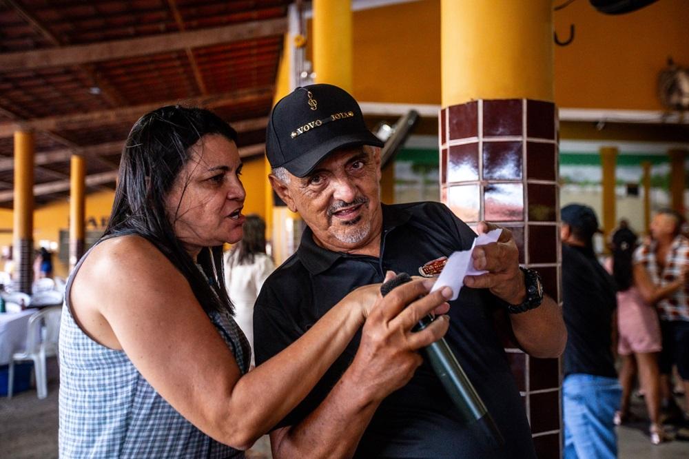 Na imagem, um close de duas pessoas, uma mulher e um homem, em um ambiente interno. O homem está no centro, com um boné preto bordado com 