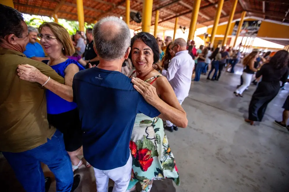 Na imagem, uma fotografia tirada em um salão de festas amplo, com teto de madeira e telhas, e pilares amarelos. O foco principal está em um casal de meia-idade ou idoso dançando. O homem está de costas para a câmera, vestindo uma camisa azul escura e calças brancas. A mulher está olhando e sorrindo para a câmera sobre o ombro do parceiro, vestindo um vestido estampado com motivos florais (branco, verde e vermelho) e tem cabelos escuros. Eles estão abraçados em posição de dança. Outros casais dançam ao redor, embaçados em segundo plano, incluindo um casal à esquerda, onde o homem veste uma camisa cáqui e a mulher uma blusa azul vibrante. O piso é de concreto. A luz natural entra pela lateral.