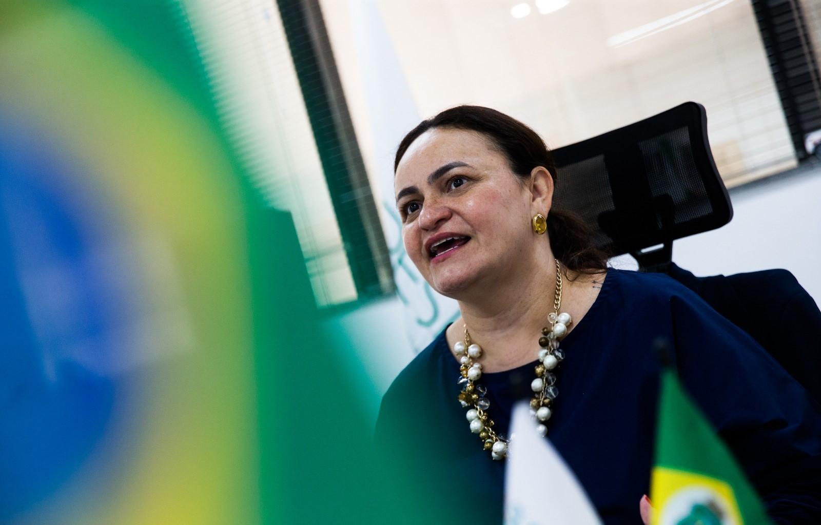 Juliana Barroso, superintendente da Supesp, falando em uma reunião, com bandeira do Brasil e fundo de ambiente de escritório.