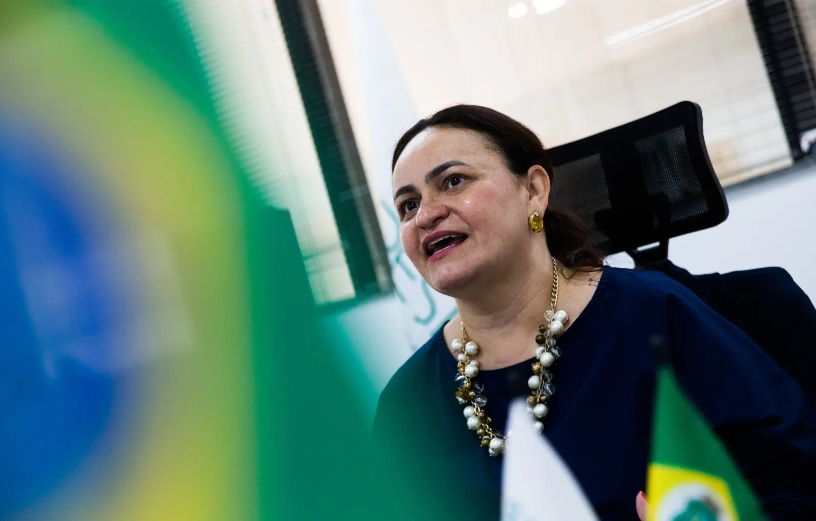 Juliana Barroso, superintendente da Supesp, falando em uma reunião, com bandeira do Brasil e fundo de ambiente de escritório.