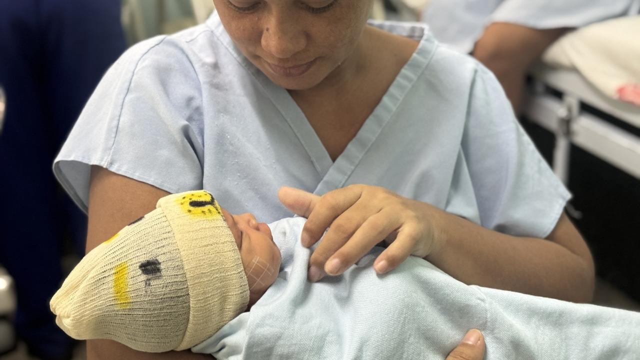 Recém-nascido em centro de parto com chapéu de leão sorrindo enquanto é segurado por sua mãe em ambiente hospitalar.