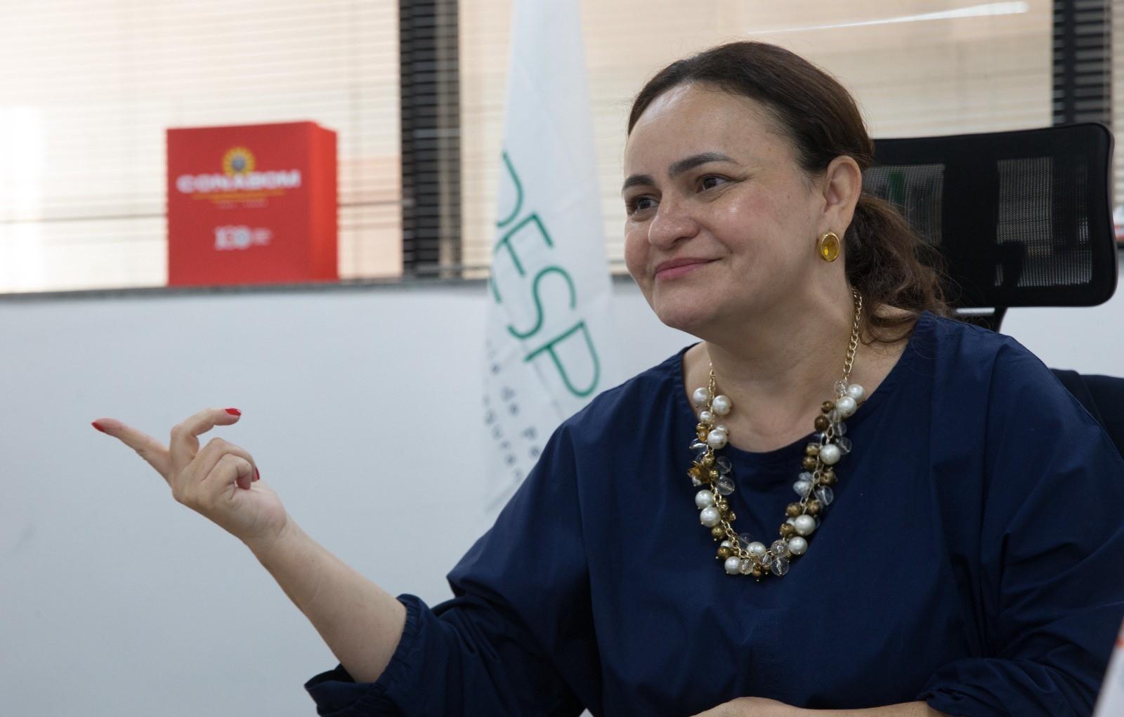 Juliana Barroso em ambiente de escritório, usando blusa azul, com acessórios de joias e sentada em cadeira de escritório, praticando postura confiante.