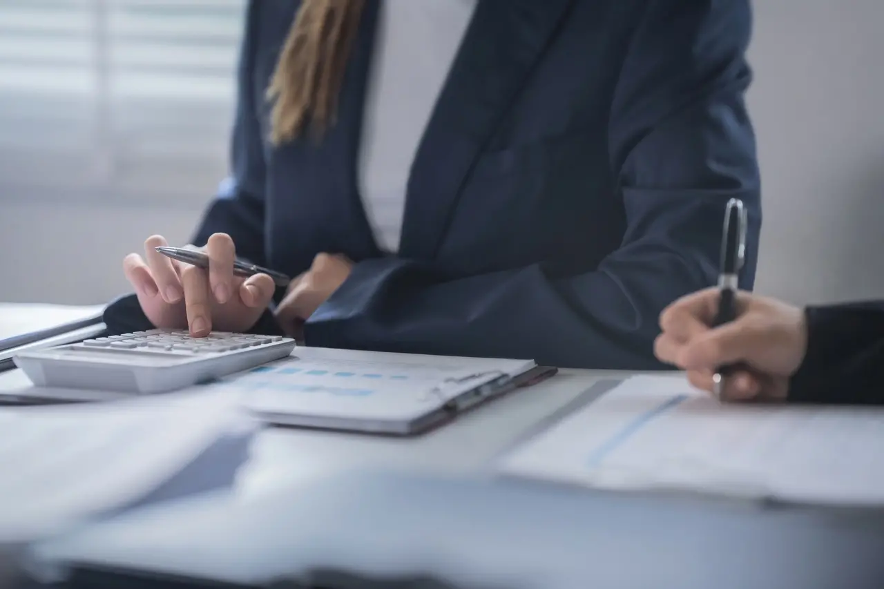Torso de pessoa de cabelo liso, loiro e longo até a altura do peitoral sentada atrás de uma mesa branca usando terno azul marinho e blusa branca. Na mesa, a mão está segurando uma caneta e usando uma calculadora branca, com anotações em uma mesa de escritório.