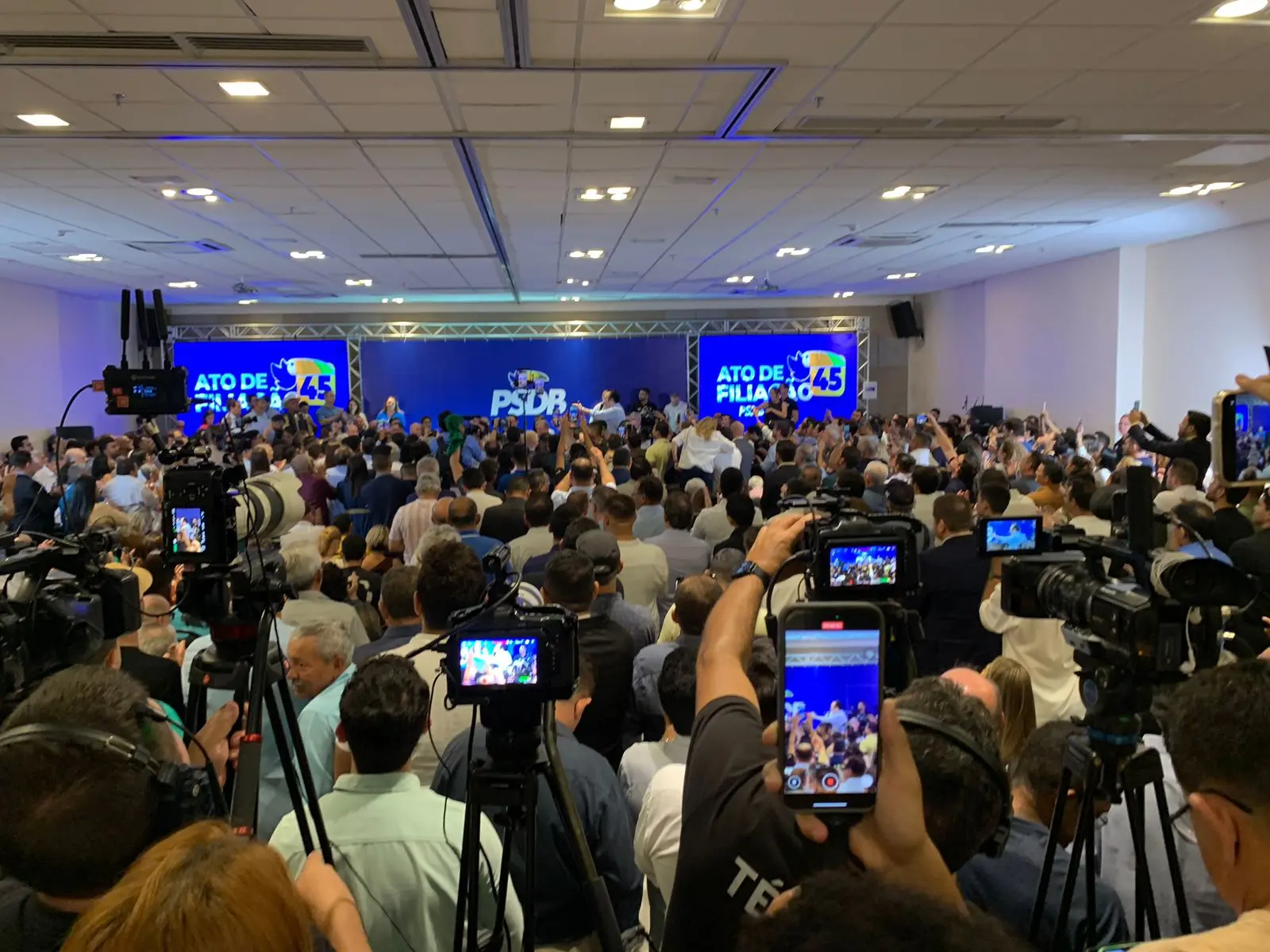 Auditório em que acontece a filiação de Ciro ao PSDB, com muitas pessoas e, ao fundo, um telão com a logomarca do partido