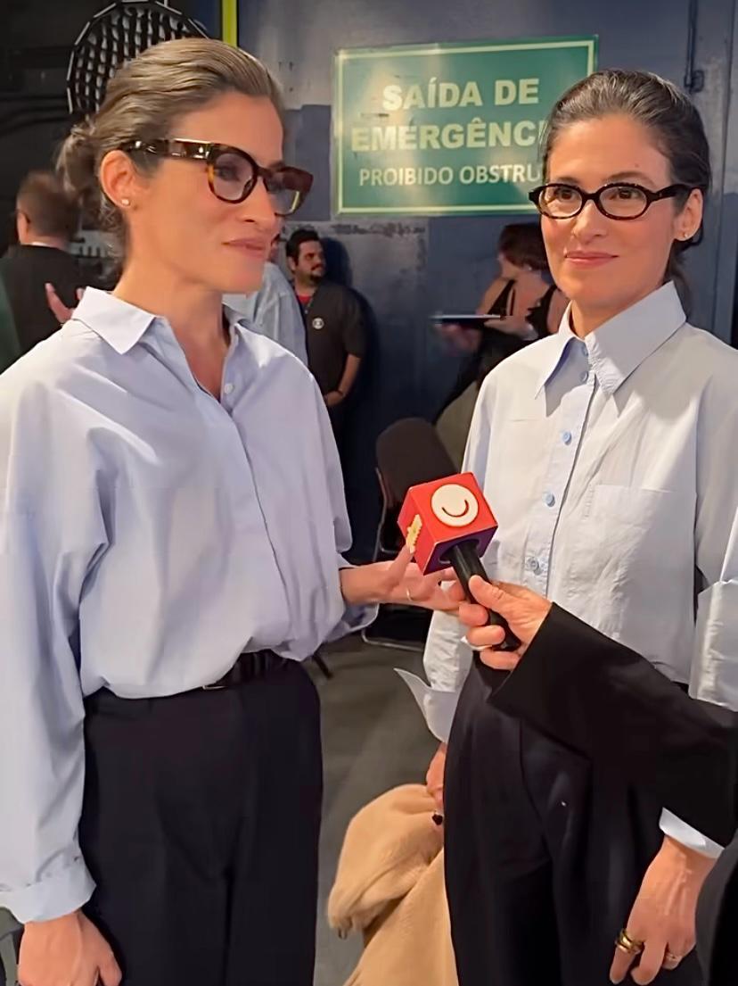 Duas mulheres usando óculos e camisas brancas em entrevista em ambiente de evento, com sinal de saída de emergência ao fundo