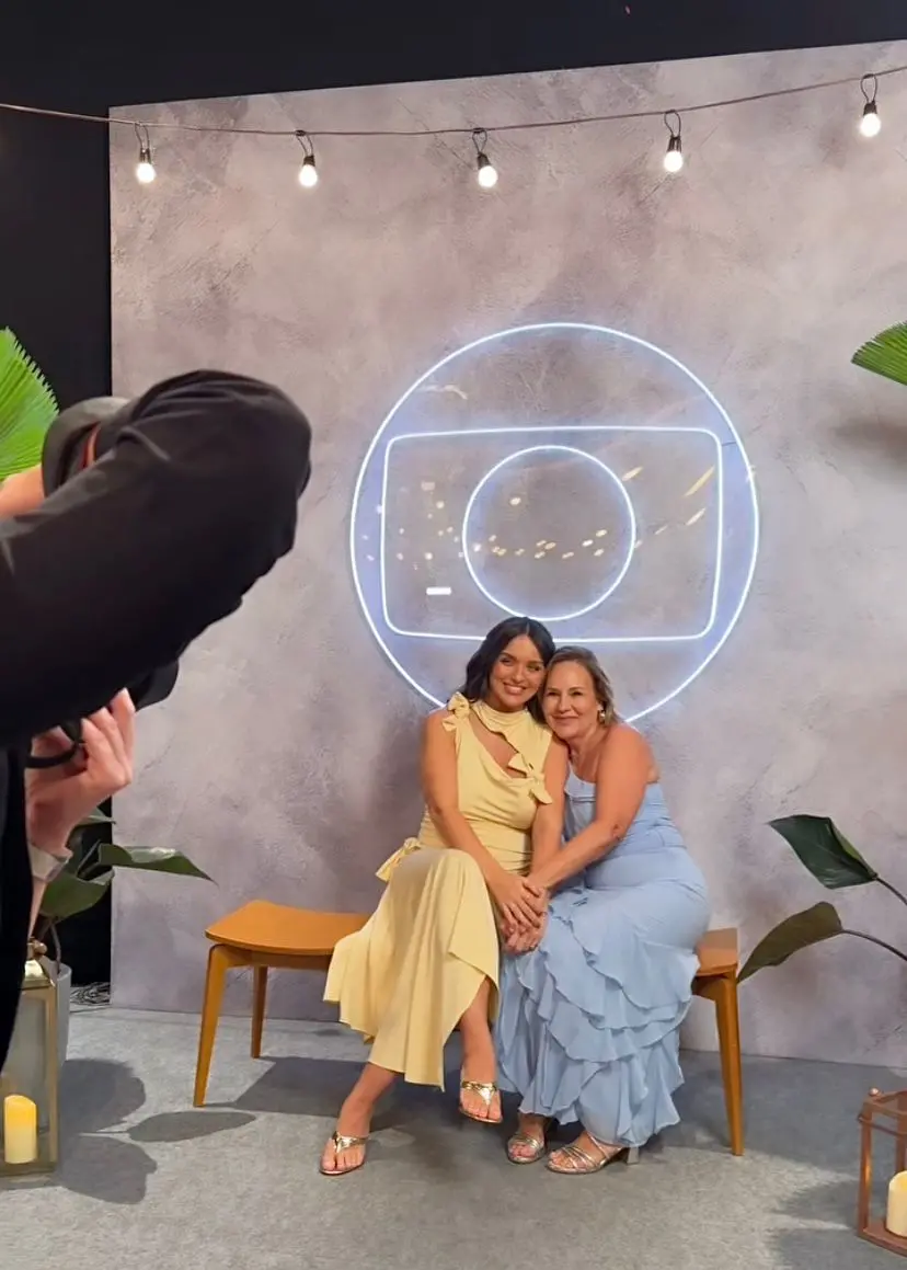 Duas mulheres sorrindo juntas para a câmera, sentadas em um banco de madeira, com fundo de parede de concreto e neon, decoradas com luzes de filamento e plantas ao redor.