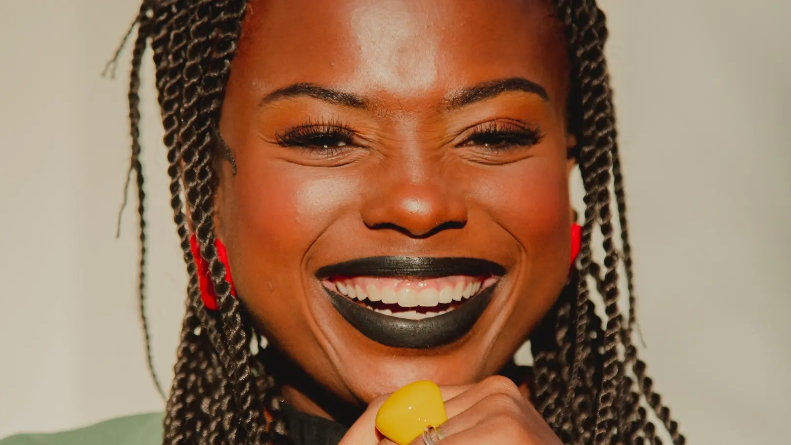 Mulher negra sorri amplamente em um retrato iluminado pela luz natural. Ela tem tranças, batom preto, brincos vermelhos e usa um anel amarelo chamativo, transmitindo alegria e confiança.