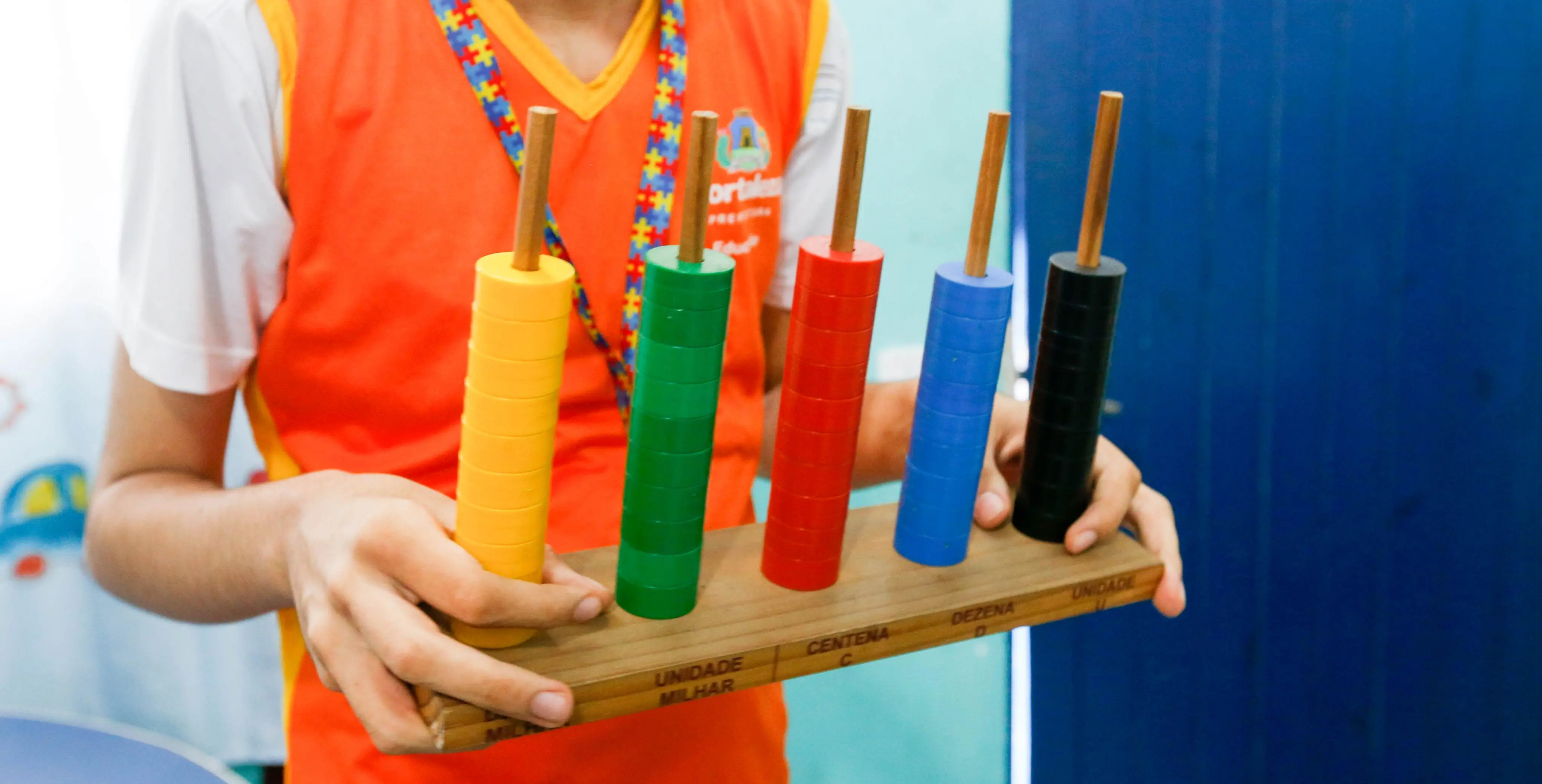 Estudante segura material pedagógico de contagem com peças coloridas empilhadas em hastes de madeira, usado para ensinar valores de unidade, dezena, centena e milhar. O aluno veste farda escolar laranja e branca com crachá estampado com peças de quebra-cabeça coloridas, símbolo da inclusão.