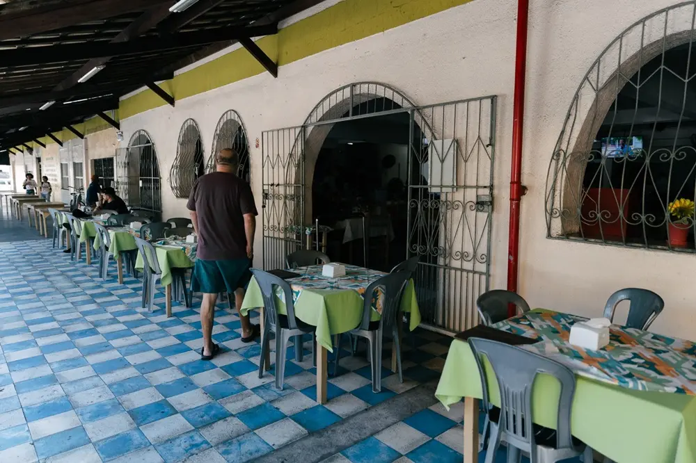 Na imagem, uma visão de uma área de refeições externa ou varanda, possivelmente de um restaurante. Há várias mesas de plástico com toalhas de mesa verde-claras e cadeiras de plástico cinza dispostas em fileiras, alinhadas ao longo de uma parede clara. O chão é um padrão de xadrez de azulejos azuis e brancos. Um homem de camiseta marrom e shorts azuis está de costas, caminhando em direção a uma abertura em arco com um portão de ferro aberto, que leva a um espaço interno escuro. Na parede, há janelas em arco com grades de ferro. O teto é inclinado e tem vigas de madeira escura.