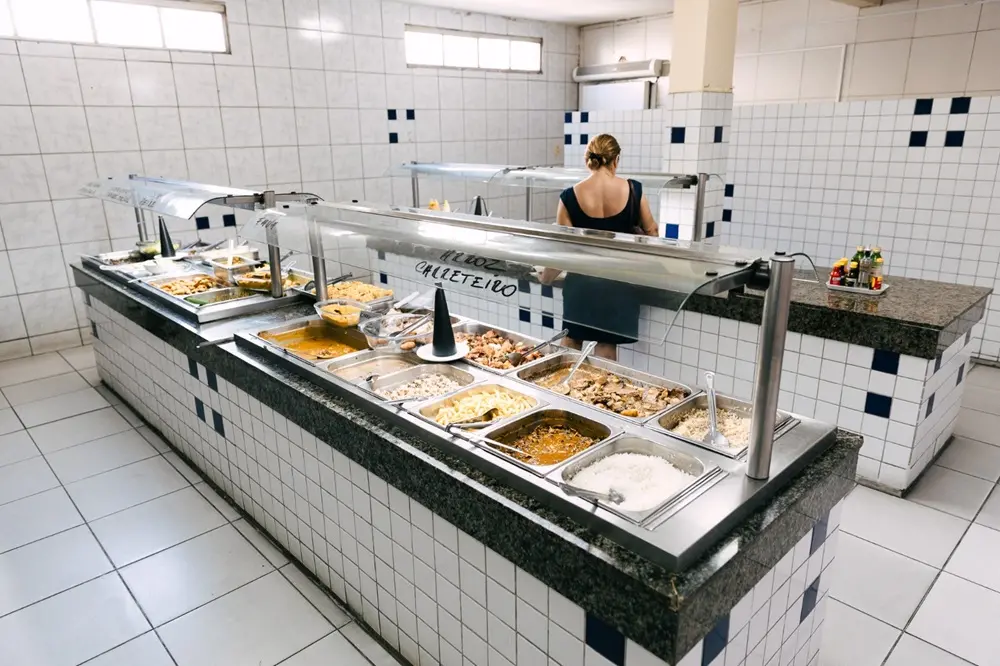 Na imagem, uma foto de uma área de buffet de um restaurante self-service. O balcão do buffet é longo e retangular, com revestimento de azulejos brancos e detalhes em azul marinho. Bandejas de metal cheias de vários pratos de comida, como arroz, feijão, e carne, estão dispostas no balcão. Um protetor de acrílico está sobre as bandejas de comida, e há um cartaz preso a ele com a palavra 