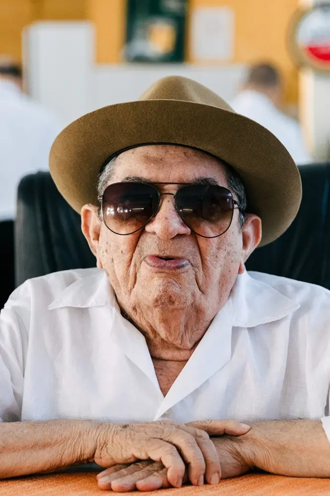 Na imagem, um close-up de um senhor idoso, de pele clara, que está sentado e olhando diretamente para a câmera. Ele veste uma camisa branca de linho de gola aberta e um chapéu Fedora marrom. Ele está usando óculos escuros grandes, com armação fina, que cobrem seus olhos. Suas mãos estão repousadas e cruzadas sobre uma superfície laranja. O fundo está desfocado, com tons de amarelo/laranja na parte superior e a silhueta de uma ou duas pessoas de camisa branca ao fundo. A iluminação é suave e natural.
