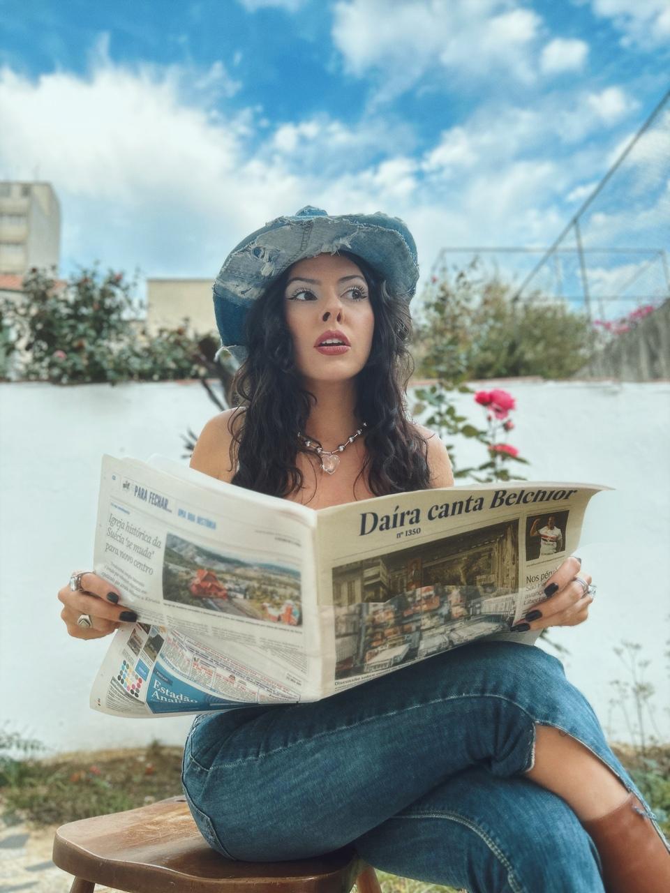 Imagem de divulgação do projeto da cantora Daíra cantando Belchior; ela aparece segurando um jornal com a manchete 'Daíra canta Belchior' e usa chapéu e calça jeans.