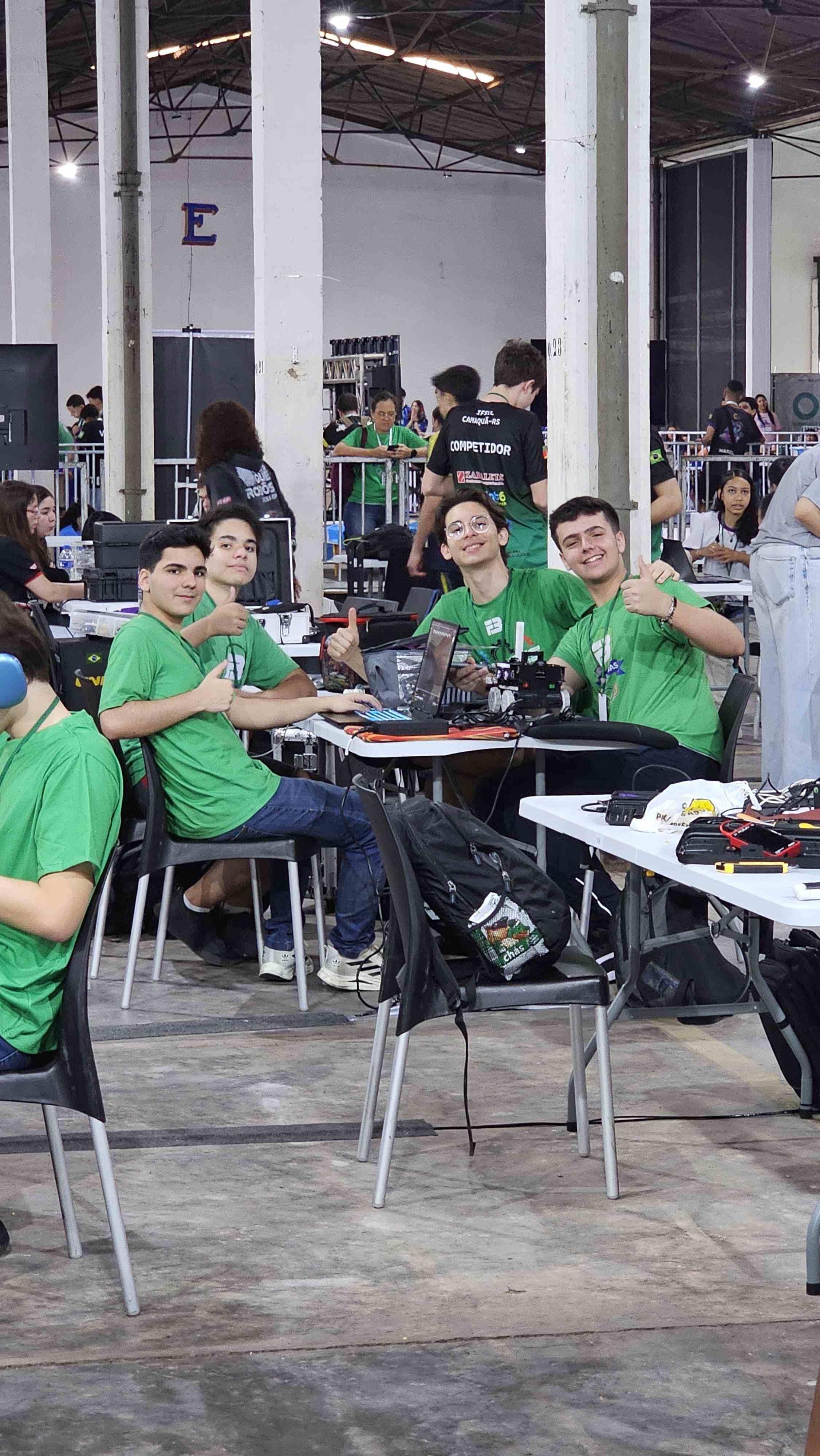 Grupo de jovens em evento de tecnologia com computadores e equipamentos de robótica, usando camisetas verdes e mostrando entusiasmo na competição.