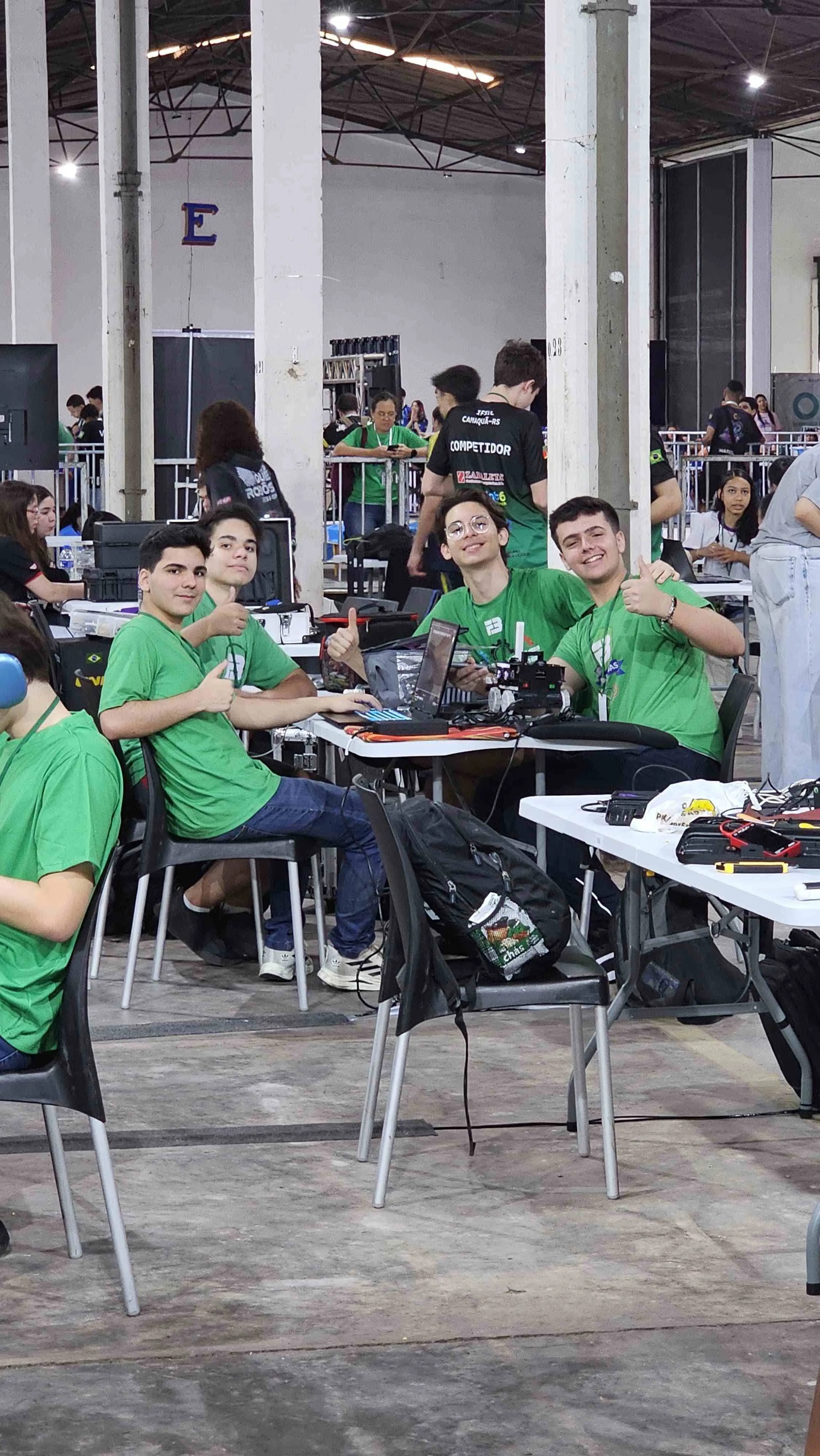 Grupo de jovens em evento de tecnologia com computadores e equipamentos de robótica, usando camisetas verdes e mostrando entusiasmo na competição.