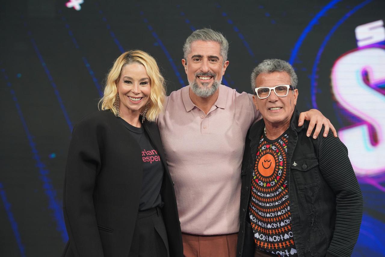 Imagem de três pessoas sorridentes posando juntas em um evento, incluindo uma mulher de cabelo louro cacheado, um homem de cabelo cinza e barba, e outro homem com óculos brancos, todos aparentando estar felizes.