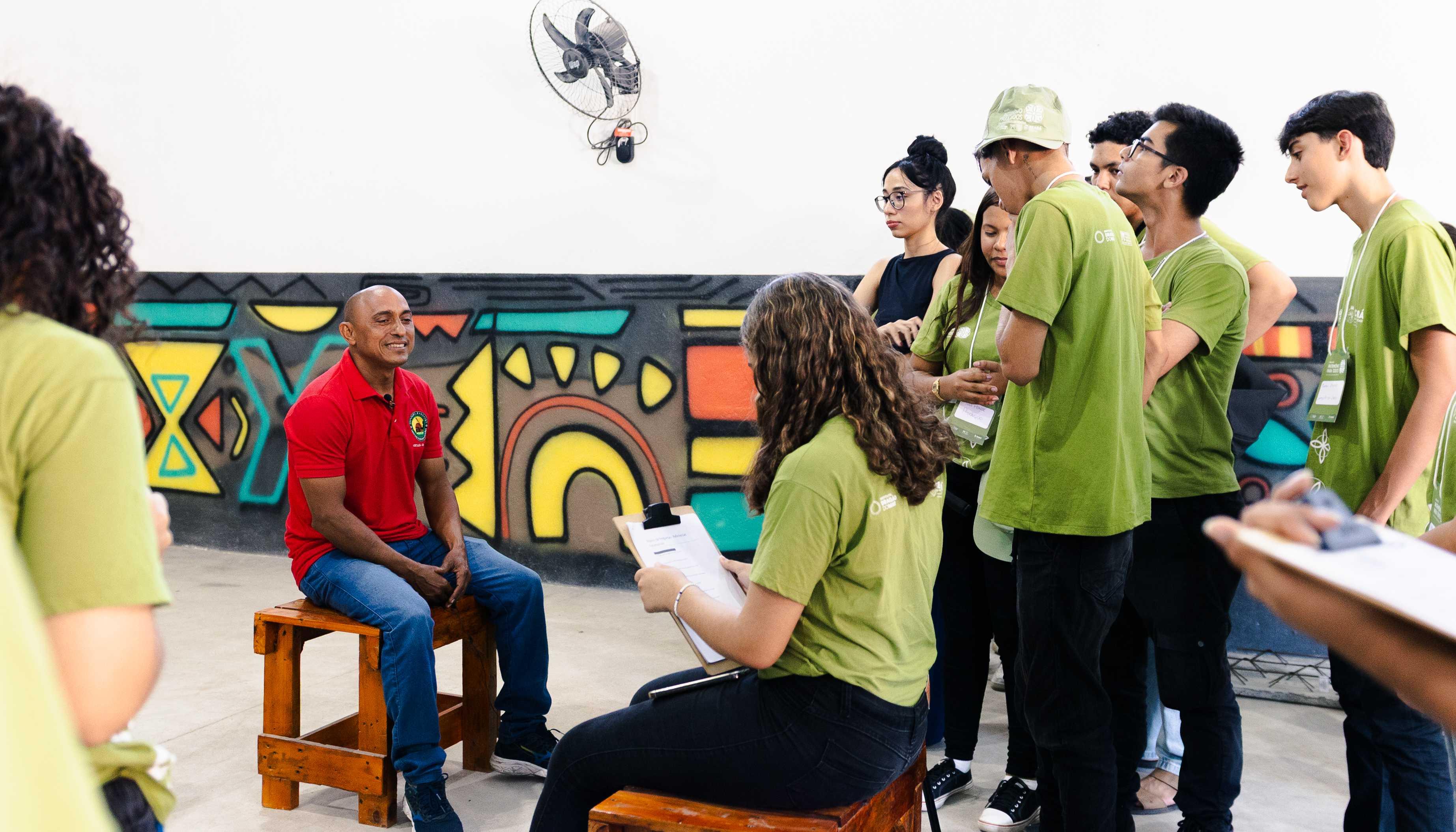 Grupo de jovens usando camisetas verdes entrevista um homem de camisa vermelha que está sentado em um banco de madeira. Eles estão em um espaço interno com parede colorida ao fundo, decorada com grafites geométricos. O clima é descontraído e educativo.