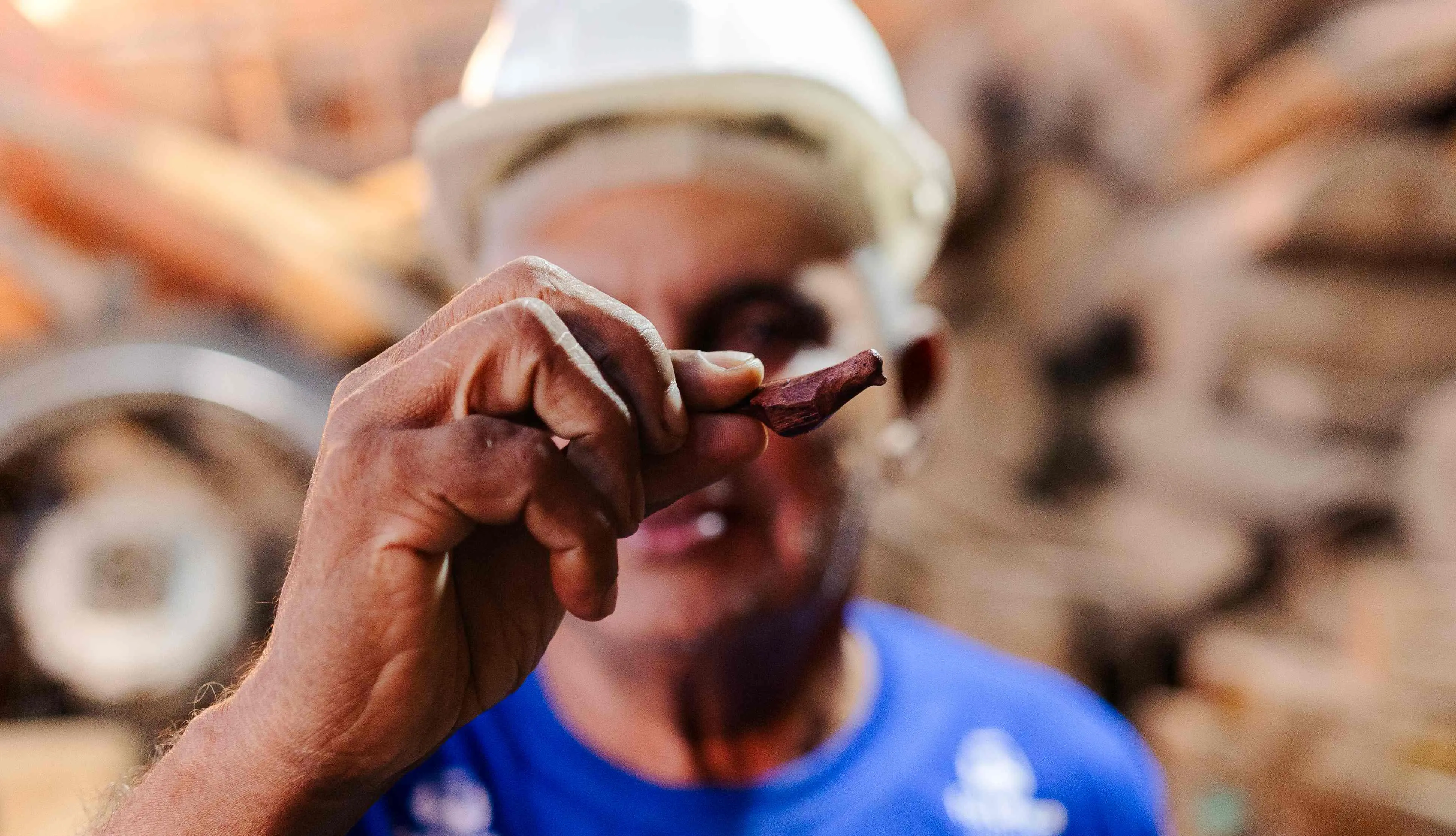 Homem usando capacete branco segura uma pequena peça de madeira esculpida em forma de pássaro, mostrando-a em primeiro plano. O fundo está desfocado, revelando um ambiente de oficina com madeira e ferramentas.