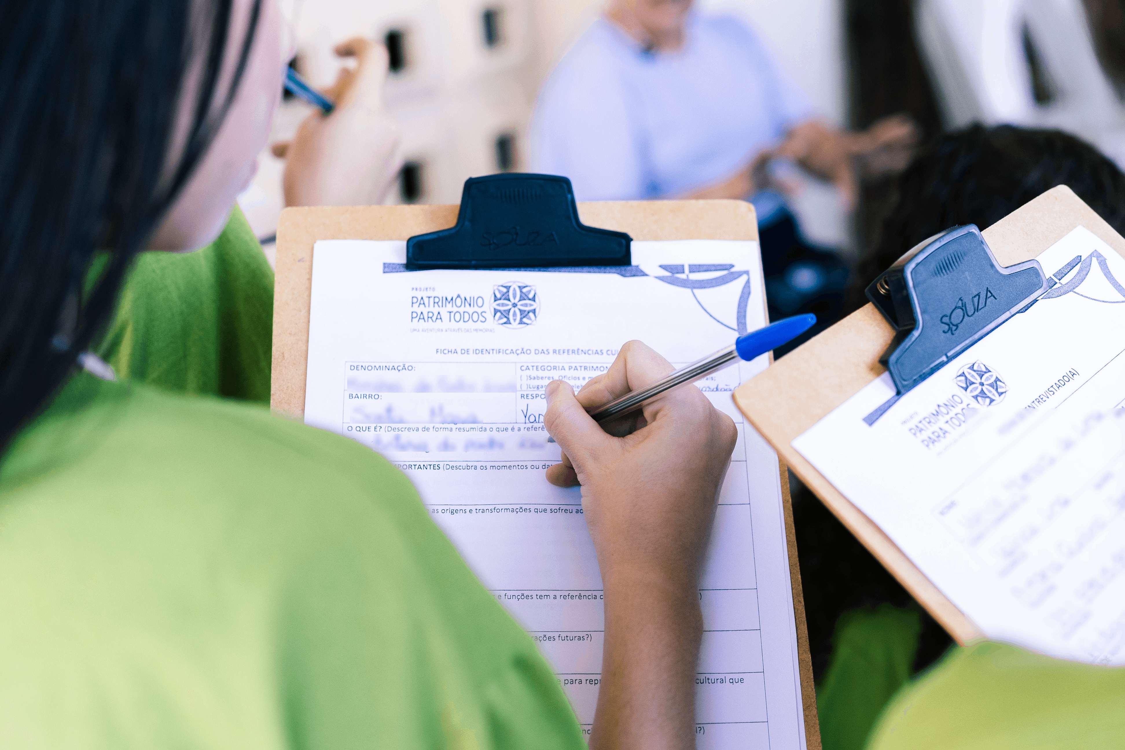 Um close-up mostra uma pessoa preenchendo um formulário de Inventário Participativo do projeto Patrimônio para Todos em uma prancheta. A pessoa está de camisa verde-limão, escrevendo dados como denominação e bairro.