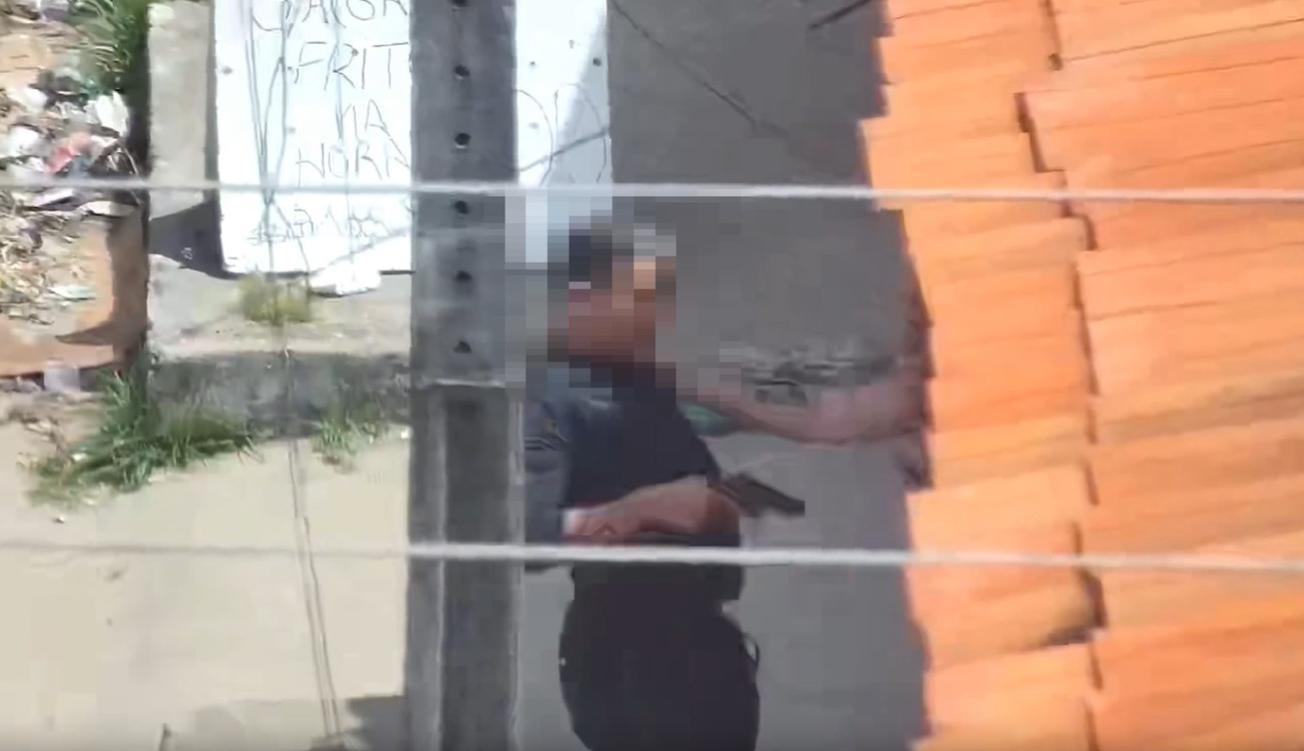 Criminoso, com arma em punho, está de pé em uma calçada próxima a um muro e a um telhado de telhas alaranjadas. A cena é vista de cima, com fios elétricos atravessando a imagem e parte de entulho visível no canto esquerdo. Ao fundo, há uma parede com uma inscrição feita à mão.