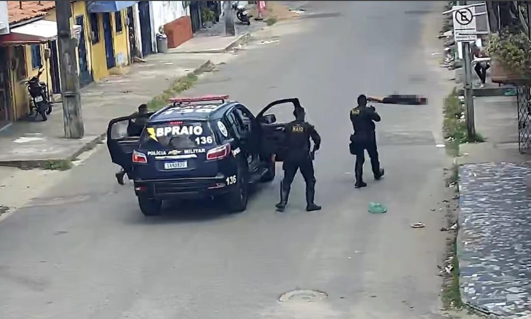 Viatura do Batalhão de Policiamento de Rondas e Ações Intensivas e Ostensivas (BPRAIO) estacionada em uma rua de bairro, com portas abertas e policiais armados próximos ao veículo. Três agentes aparecem em posição de ação, enquanto uma pessoa está caída ao chão mais à frente. A cena ocorre em via asfaltada, com casas e comércios simples nas laterais.