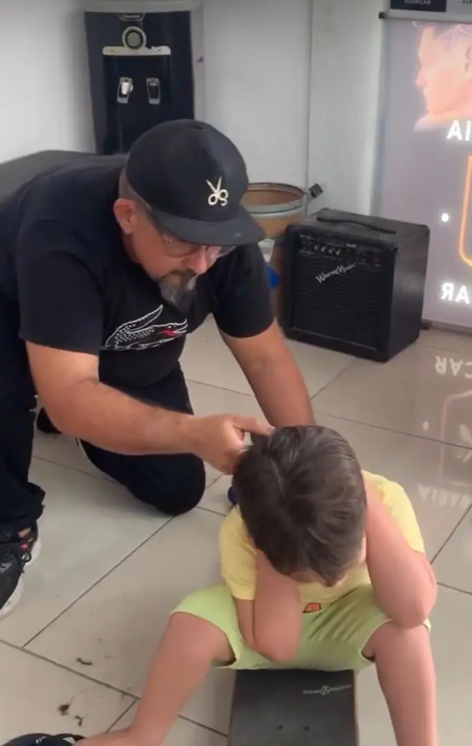 Na imagem, uma barbearia ou salão de cabeleireiro onde um homem barbudo com um boné preto e uma camisa polo preta está ajoelhado, aparentemente arrumando o cabelo de uma criança. A criança, vestindo uma camisa amarela e shorts verde-limão, está sentada no chão (ou possivelmente em um skate no chão) e cobre as orelhas com as mãos, talvez em reação ao corte de cabelo ou ao barulho. O homem está segurando uma tesoura ou uma máquina pequena perto da cabeça da criança. Ao fundo, é possível ver um bebedouro ou purificador de água escuro, um pequeno amplificador preto no chão e um cartaz ou banner na parede. O piso é de cerâmica clara.
