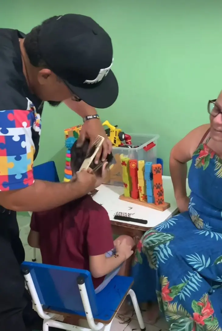 Na imagem, uma foto tirada em um ambiente que parece ser uma área de corte de cabelo para crianças ou um espaço adaptado, com uma parede verde-clara ao fundo. Um barbeiro ou cabeleireiro barbudo, usando um boné preto e uma camisa com mangas coloridas de estampa de quebra-cabeça, está cortando o cabelo de uma criança sentada em uma pequena cadeira azul. O homem está usando um pente para prender o cabelo da criança. No lado direito da foto, uma mulher de óculos, vestindo um vestido floral azul, está sentada, sorrindo e observando. Sobre uma pequena mesa branca, há brinquedos coloridos, incluindo um brinquedo de encaixe de madeira e outros blocos de construção em uma caixa. O ambiente é acolhedor e focado no atendimento infantil.