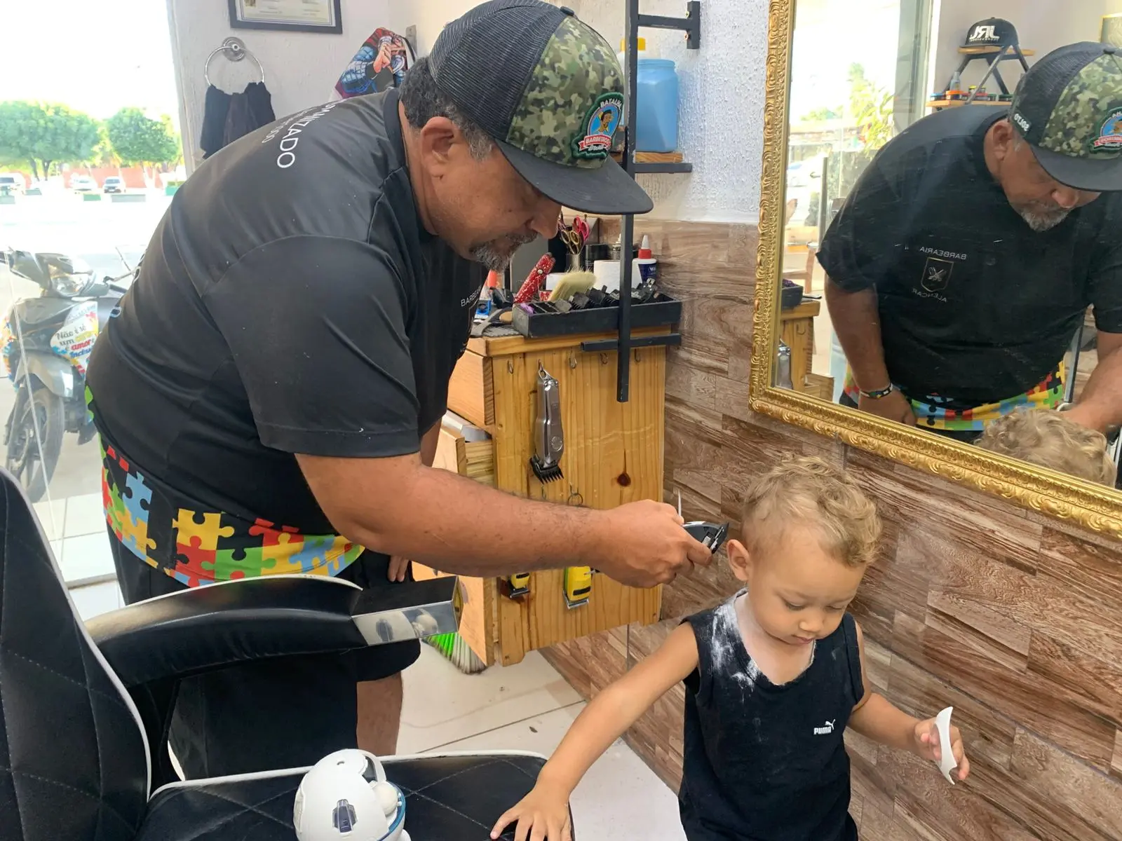 Na imagem, uma foto tirada em uma barbearia ou salão de cabeleireiro. Um barbeiro barbudo, vestindo uma camiseta preta, um avental com estampa colorida de quebra-cabeça e um boné camuflado com um emblema, está cortando o cabelo de uma criança pequena e loira, que veste uma regata preta. O barbeiro está usando uma máquina de cortar cabelo perto da orelha da criança. O cabelo e a nuca da criança estão cobertos com talco de corte de cabelo. A criança está de pé, parcialmente virada para a cadeira de barbeiro preta. Ao fundo, há um espelho grande com moldura dourada refletindo o barbeiro e o interior da loja. No reflexo, o barbeiro está usando o avental de quebra-cabeça. Móveis de madeira e o que parece ser uma motocicleta estacionada do lado de fora da loja são visíveis.