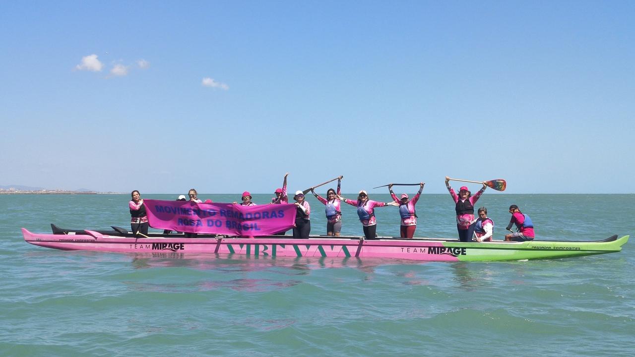 Imagem mostra grupo de sobrevivente de câncer de mama que praticam canoagem no mar de Fortaleza em pé em canoa, no mar, segurando faixa que diz movimento remadoras rosa do Brasil, em referência aos projetos de canoagem que reúnem sobreviventes da doença pelo Brasil para praticar o esporte.
