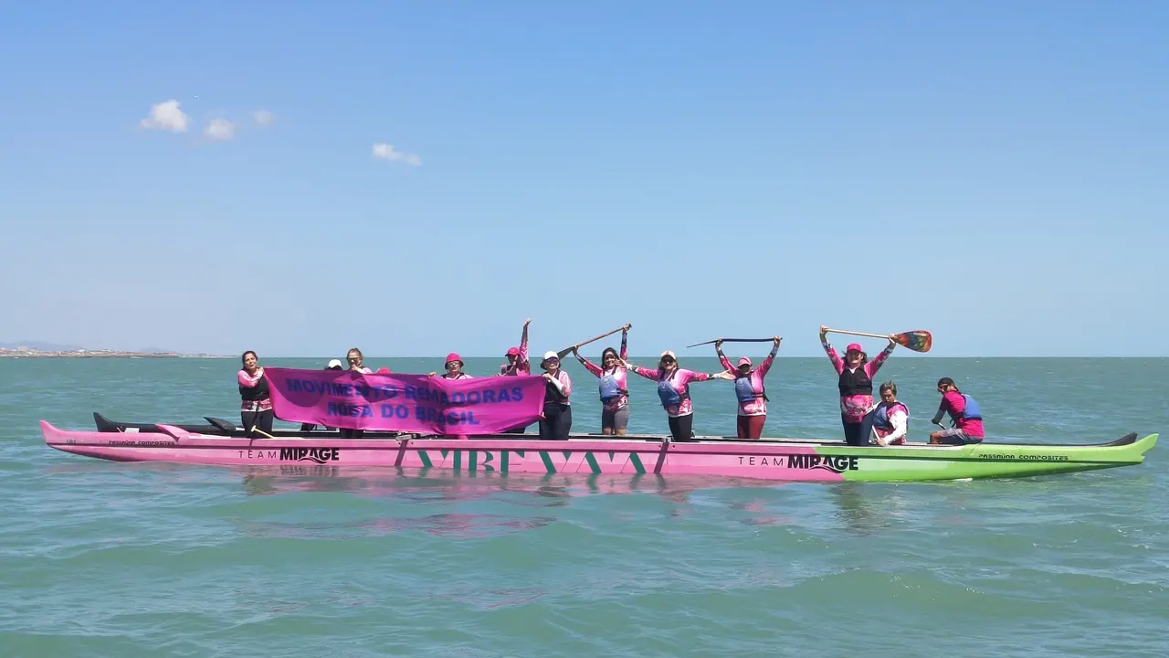 Imagem mostra grupo de sobrevivente de câncer de mama que praticam canoagem no mar de Fortaleza em pé em canoa, no mar, segurando faixa que diz movimento remadoras rosa do Brasil, em referência aos projetos de canoagem que reúnem sobreviventes da doença pelo Brasil para praticar o esporte.