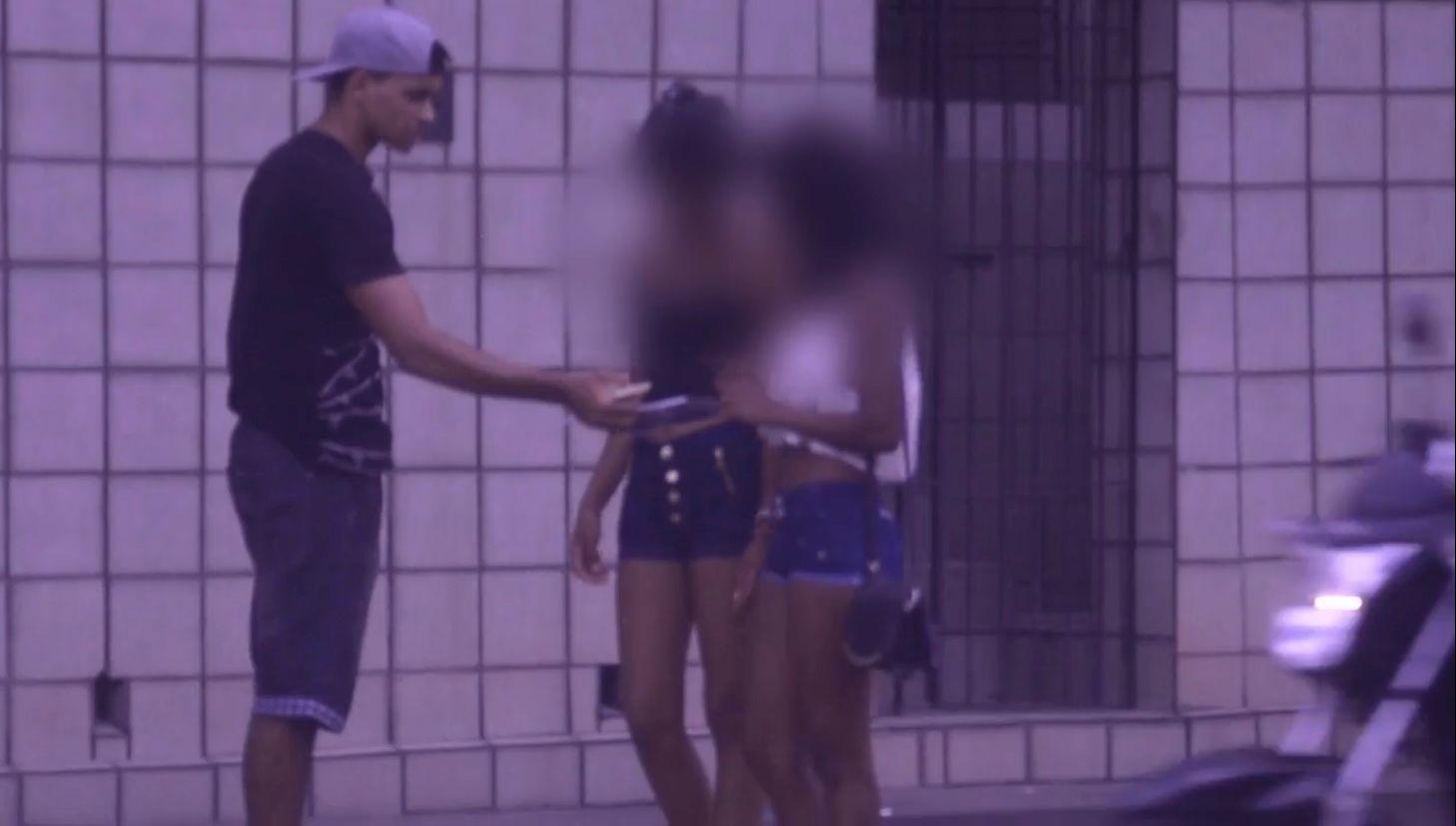 Imagem de um homem entregando uma ratoeira a uma jovem ao lado de outras duas garotas, com uma parede de tijolos ao fundo, cena noturna