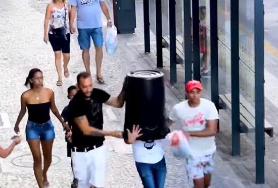 Grupo de pessoas caminhando na rua, incluindo dois homens colocando um balde reto na cabeça de um outro homem, durante o dia, com arquiteturas modernas ao fundo, em um ambiente urbano.