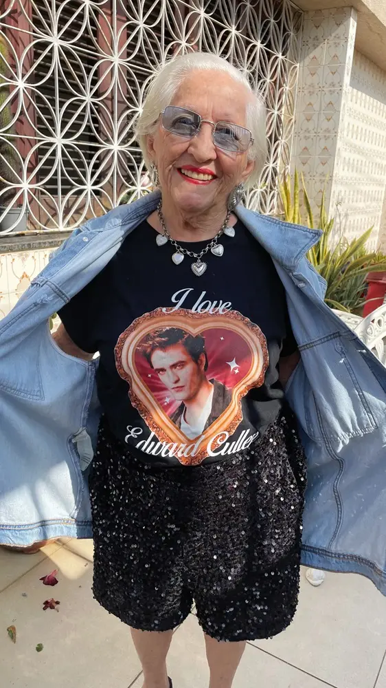 Na imagem, uma foto em close de Dona Lourdinha Reis, a fã sênior da saga Crepúsculo, sorrindo, vestindo uma roupa moderna e casual em um dia ensolarado ao ar livre. Ela tem cabelos brancos curtos e óculos de sol com lentes azuis claras. Ela está vestindo uma camiseta preta com a imagem de Edward Cullen (interpretado pelo ator Robert Pattinson) em uma moldura de coração laranja. Acima da imagem, lê-se 