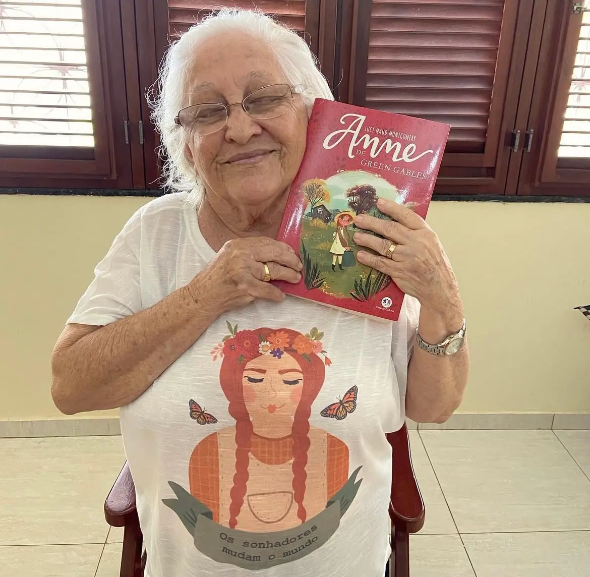 Na imagem, uma foto em close de Dona Lourdinha Reis, de pele clara e cabelos brancos, sentada em uma cadeira de madeira em um ambiente interno. Ela está usando óculos de grau e tem uma expressão serena e um leve sorriso. Ela abraça um livro de capa vermelha com a ilustração de uma jovem ruiva no campo. O título do livro é Anne de Green Gables, de Lucy Maud Montgomery. Ela está vestindo uma camiseta branca com uma estampa centralizada de uma mulher de cabelo ruivo com tranças e uma coroa de flores. Abaixo da imagem, um banner diz: 