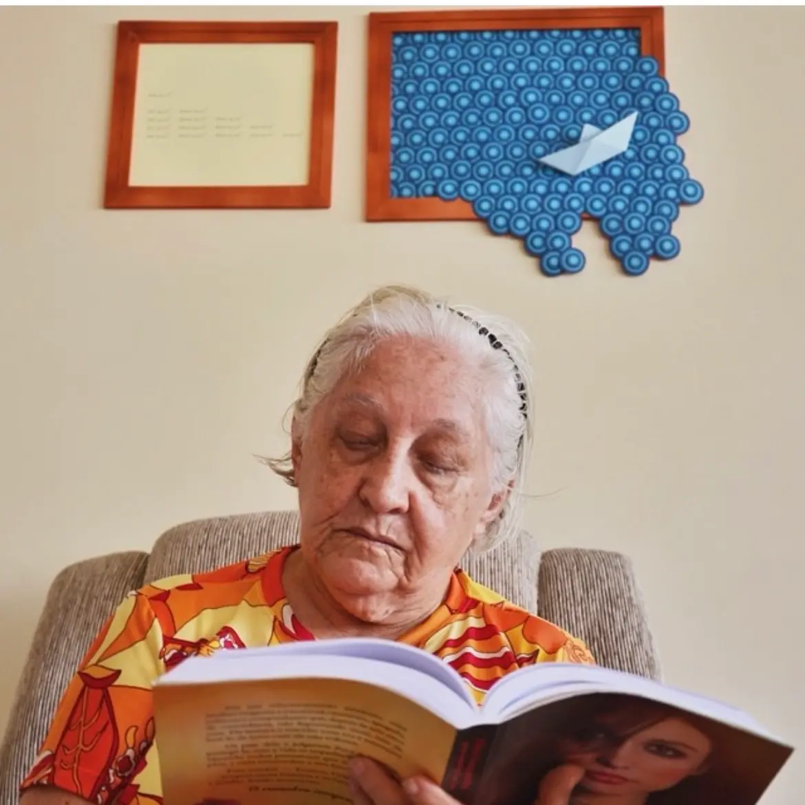 Na imagem, uma foto de close de Dona Lourdinha Reis, de pele clara e cabelos brancos puxados para trás com uma tiara preta, concentrada na leitura de um livro. Ela está sentada em uma poltrona estofada e veste uma blusa com estampa abstrata nas cores laranja e amarelo. O livro está aberto em primeiro plano, com parte de uma imagem de uma mulher ruiva na capa visível. Ao fundo, em uma parede de cor creme, há duas peças de arte emolduradas: à esquerda, um quadro simples de tom claro com escrita sutil; à direita, um quadro de madeira mais grosso com uma decoração em azul escuro e um barquinho de origami branco.