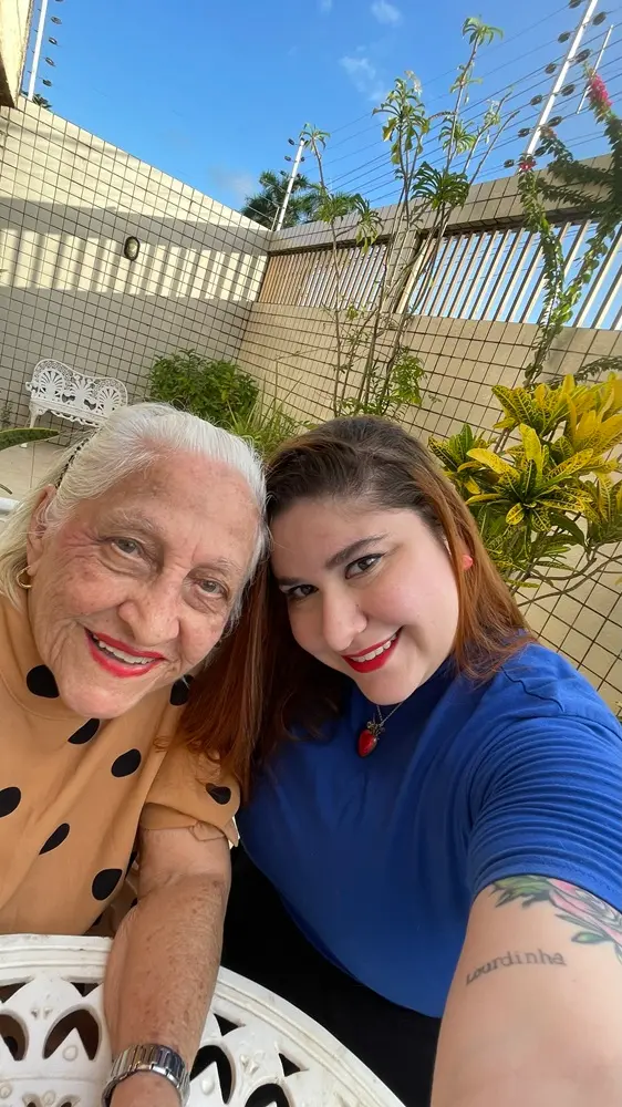 Na imagem, uma selfie vertical em que Dona Lourdinha Reis e uma mulher mais jovem estão lado a lado, sorrindo para a câmera em um dia ensolarado. Dona Lourdinha, de pele clara e cabelo branco, usa uma blusa marrom claro com bolinhas pretas e um batom vermelho vibrante. Ela também está usando um relógio de pulso prateado e brincos pequenos. A mulher mais jovem tem cabelo castanho-avermelhado e está usando uma camiseta azul vibrante e batom vermelho. Ela tem uma tatuagem no braço esquerdo que parece dizer 