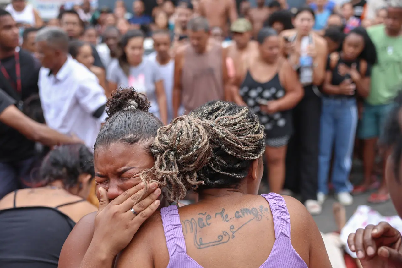 Mulher chorando em protesto ou reunião pública com uma multidão ao fundo, mostrando dor emocional, com destaque para a tatuagem nas costas e cabelo trançado.