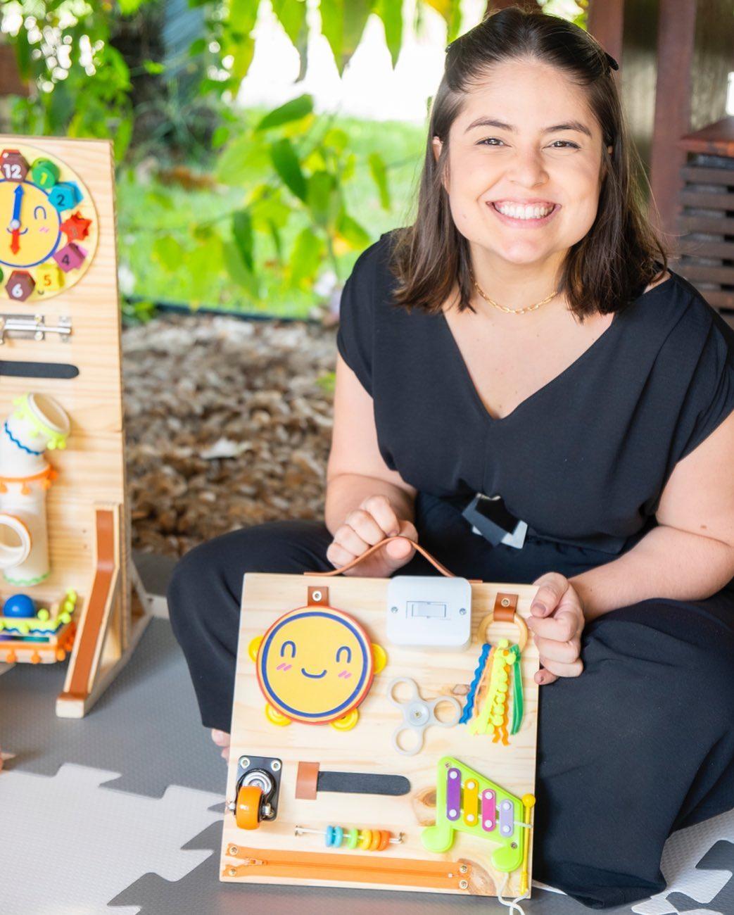 paula segurando painel de madeira com itens que transformam a madeira em um painel sensorial. ela veste roupa preta e sorri.