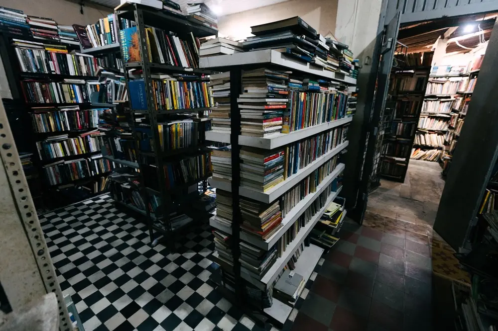 Na imagem, uma visão de dentro de um sebo (livraria de livros usados), focando nas estantes abarrotadas de livros. Em primeiro plano, no lado esquerdo, o piso é de ladrilho xadrez preto e branco. No centro, uma estante modular de livros, empilhados tanto nas prateleiras quanto no topo, serve como um divisor de ambientes, separando a área do piso xadrez de um corredor à direita. À esquerda e atrás dessa estante central, há mais estantes altas de metal, repletas de livros. À direita, uma porta ou abertura de corredor revela uma área com o piso em tons de xadrez marrom e um conjunto mais profundo de estantes escuras e ainda mais livros. A iluminação é difusa, mas suficiente para destacar a densidade do inventário. A imagem transmite a sensação de um local muito cheio e dedicado aos livros.