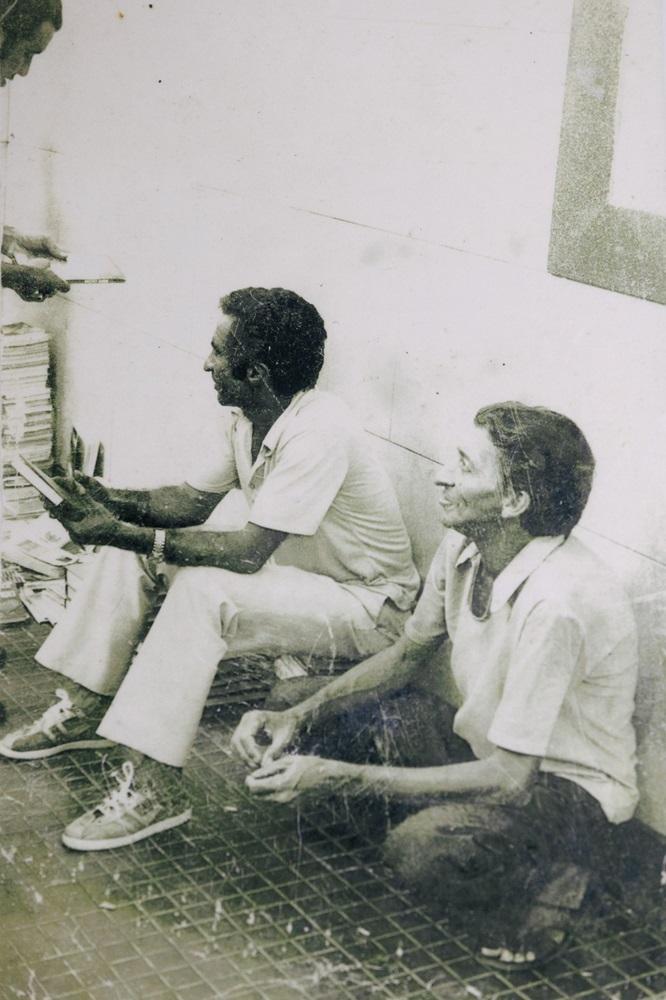 Na imagem, uma fotografia antiga, em preto e branco (ou sépia desbotada), que mostra duas pessoas sentadas no chão. A foto tem arranhões e marcas de desgaste. O homem à esquerda está sentado com as pernas esticadas, vestindo calças claras e uma camisa polo clara, e tênis. Ele segura o que parece ser um livro ou papéis e olha para a esquerda, fora do enquadramento. A mulher à direita está sentada, possivelmente ajoelhada, mais próxima da câmera, também vestindo uma camisa clara. Ela sorri, olhando para a direita, também para fora do enquadramento. Eles estão encostados em uma parede clara, e o chão é escuro, com um padrão quadriculado ou gradeado. No canto superior esquerdo, parte de uma terceira pessoa e uma pilha de papéis ou livros são visíveis.