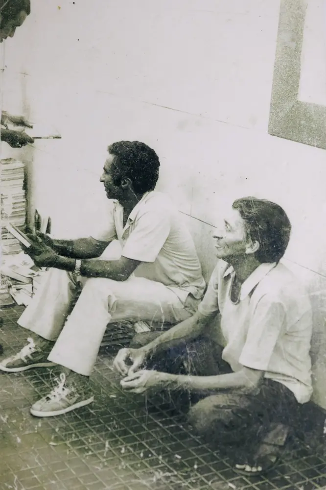 Na imagem, uma fotografia antiga, em preto e branco (ou sépia desbotada), que mostra duas pessoas sentadas no chão. A foto tem arranhões e marcas de desgaste. O homem à esquerda está sentado com as pernas esticadas, vestindo calças claras e uma camisa polo clara, e tênis. Ele segura o que parece ser um livro ou papéis e olha para a esquerda, fora do enquadramento. A mulher à direita está sentada, possivelmente ajoelhada, mais próxima da câmera, também vestindo uma camisa clara. Ela sorri, olhando para a direita, também para fora do enquadramento. Eles estão encostados em uma parede clara, e o chão é escuro, com um padrão quadriculado ou gradeado. No canto superior esquerdo, parte de uma terceira pessoa e uma pilha de papéis ou livros são visíveis.