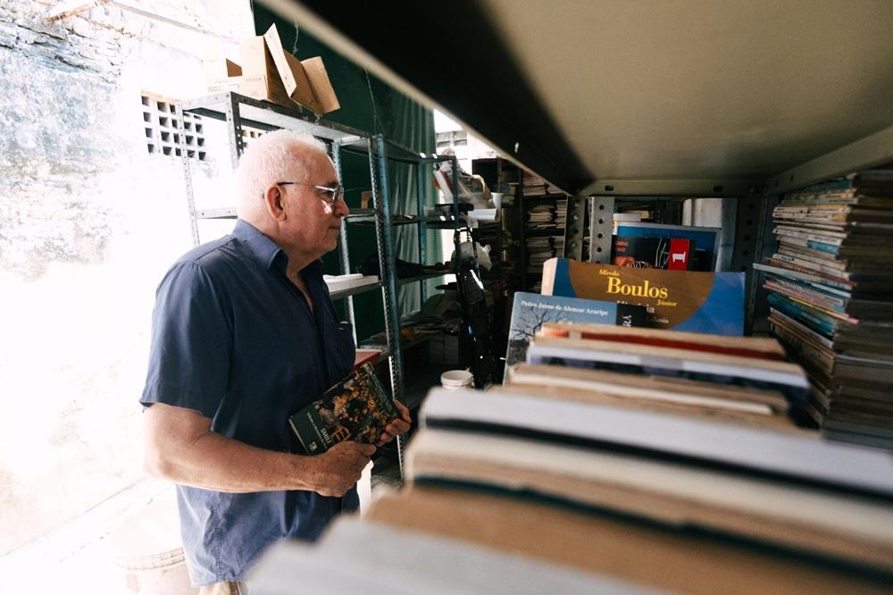 Na imagem, um homem idoso, de pele clara, cabelos brancos curtos e óculos, está em pé dentro de uma livraria de livros usados (sebo), segurando um livro com a capa estampada nas mãos. Ele veste uma camisa de botão azul escura de manga curta e está posicionado de perfil, olhando para a esquerda, para fora do enquadramento. A foto é cortada pela perspectiva, com uma prateleira ou mesa escura em primeiro plano no lado direito, onde pilhas de livros se encontram. Atrás do homem, à esquerda, há uma parede texturizada, possivelmente antiga, com uma ventilação em treliça. Mais prateleiras de metal cheias de livros e objetos são visíveis ao fundo. A luz natural forte vem do lado esquerdo, iluminando bem o rosto e a camisa do homem, criando um contraste com as áreas mais escuras no lado direito e superior.